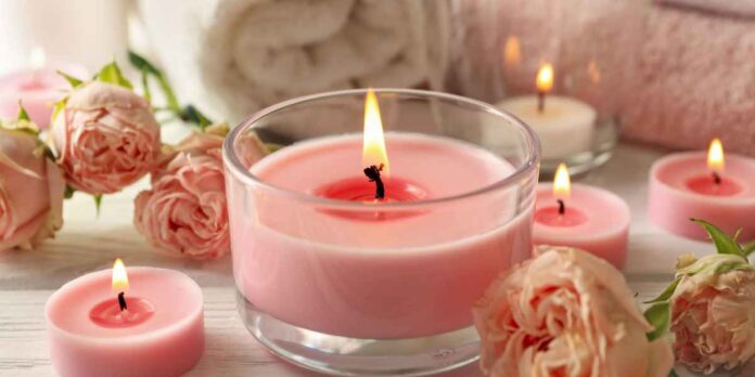 Velas aromáticas em pote de vidro aceso, cercada por velas menores, rosas cor-de-rosa e toalha, em fundo de madeira clara.