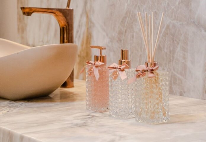 Imagem de uma pia de banheiro elegante com uma torneira de cobre e uma bancada de mármore. Sobre a bancada, há dois aromatizantes de ambiente em frascos de vidro com design cristalino, decorados com laços cor-de-rosa e varetas de difusão, ao lado de um dispensador de sabonete líquido com detalhes em cobre.